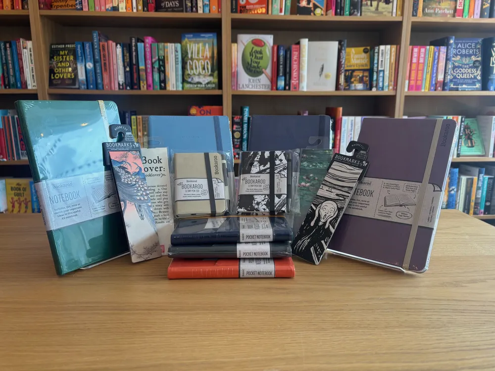 Notebooks in green, blue and plum, pocket notebooks and bookmarks arranged on a table in front of the book shelves